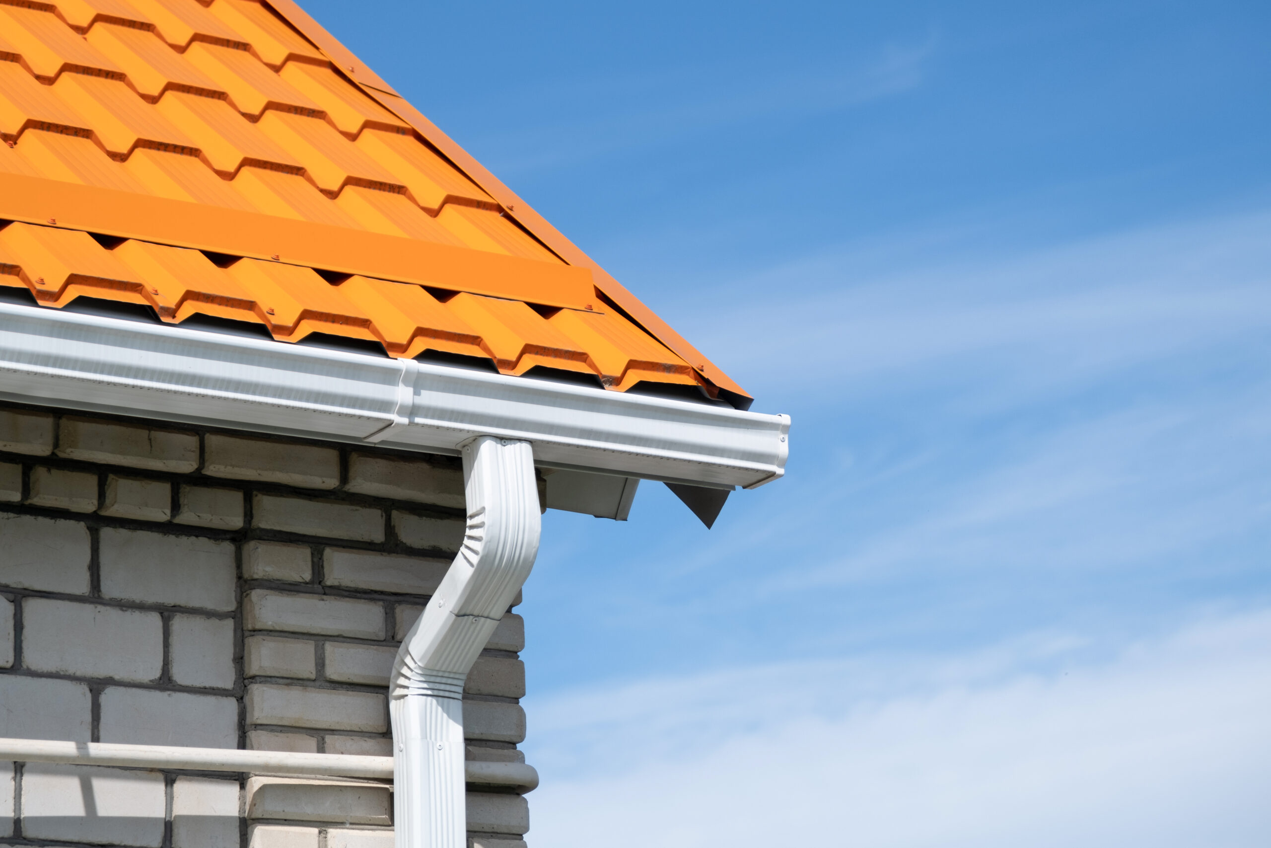 Corner of a brick house showing the roof edge, white gutter and downspout where the roof plane meets the wall